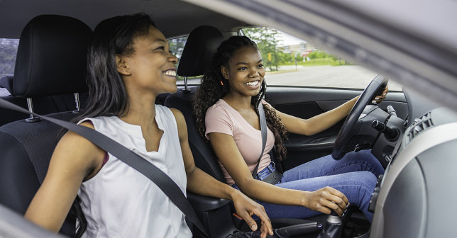 two teenagers smiling, wearing seatbelts and driving a vehicle